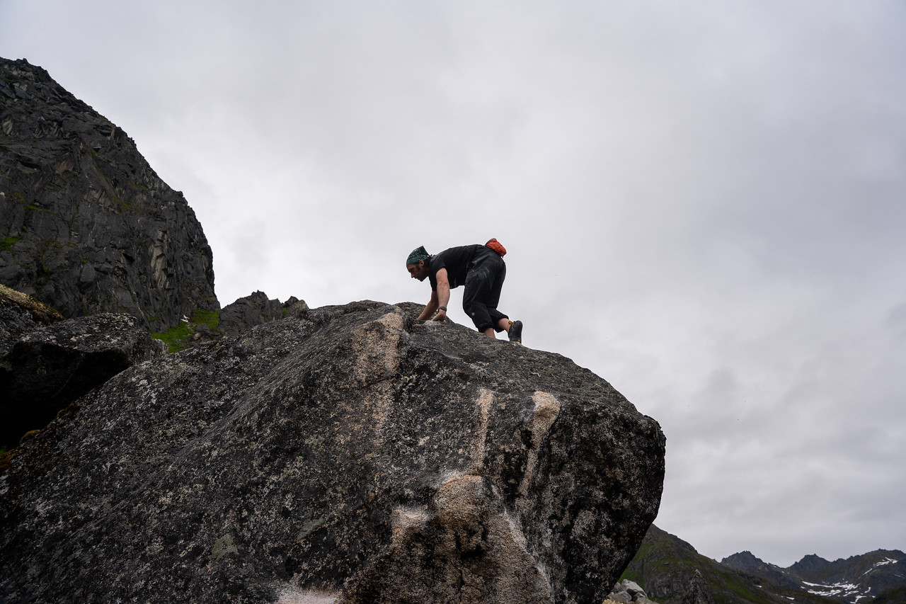 Climber finishes a boulder problem, crawling carefully over the top of a rounded rock formation under an overcast sky. Jagged cliffs and alpine vegetation fill the background.