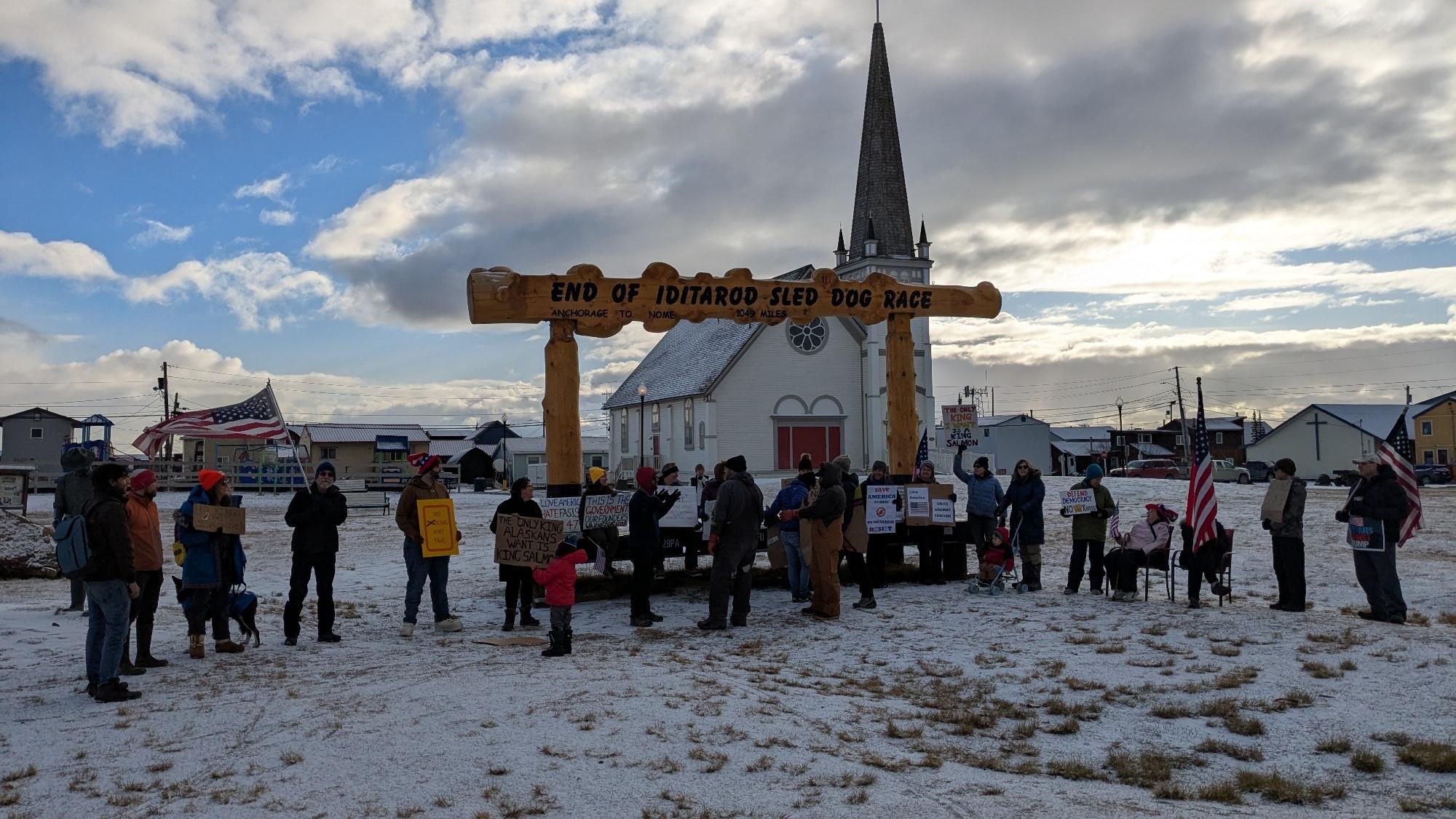 A group of people gathered in front of the Iditarod arch in Nome, with a background of the community center known as Old St. Joe's (used to be catholic church) and a partly cloudy sky. A light skiff of snow covers the ground.