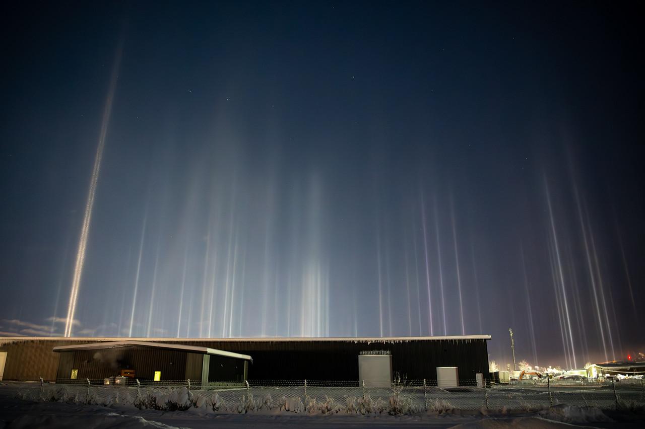 Light pillars above an industrial area in Fairbanks, Alaska at night. A long, low industrial building sits in the foreground behind a chain-link fence, surrounded by snow. Numerous vertical light pillars rise from bright lights around the buildings, forming white and faintly colored columns against a dark blue sky. A single brighter pillar stands out on the left side of the frame.