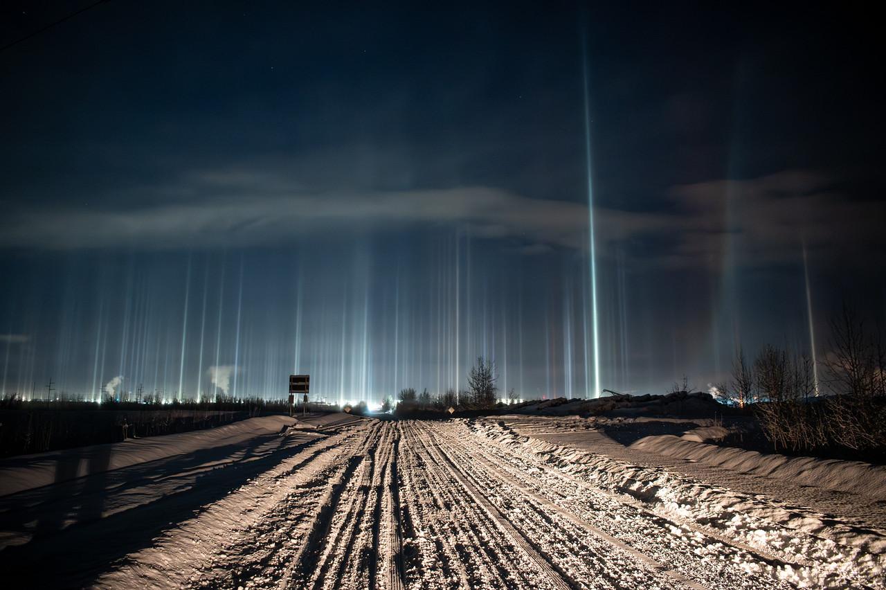 Nighttime winter scene in Fairbanks, Alaska. A snow-covered road with deep tire tracks runs straight into the distance. Beyond the road, bright city lights create numerous tall, vertical light pillars rising into the dark sky. The pillars vary in brightness and height, forming pale white and bluish columns above the horizon beneath a low cloud layer.