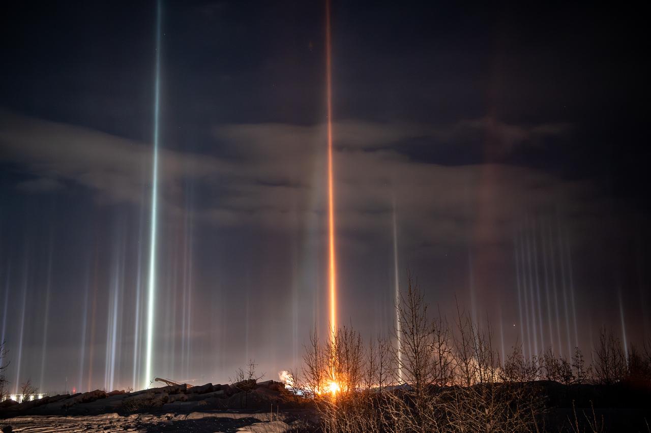 Vertical light pillars above a snowy landscape in Fairbanks, Alaska at night. A bright orange light source near the ground produces a vivid orange pillar extending upward, surrounded by multiple white and bluish pillars from nearby lights. Leafless shrubs and small trees appear in silhouette in the foreground, with snowbanks and a low ridge separating the viewer from the lights.