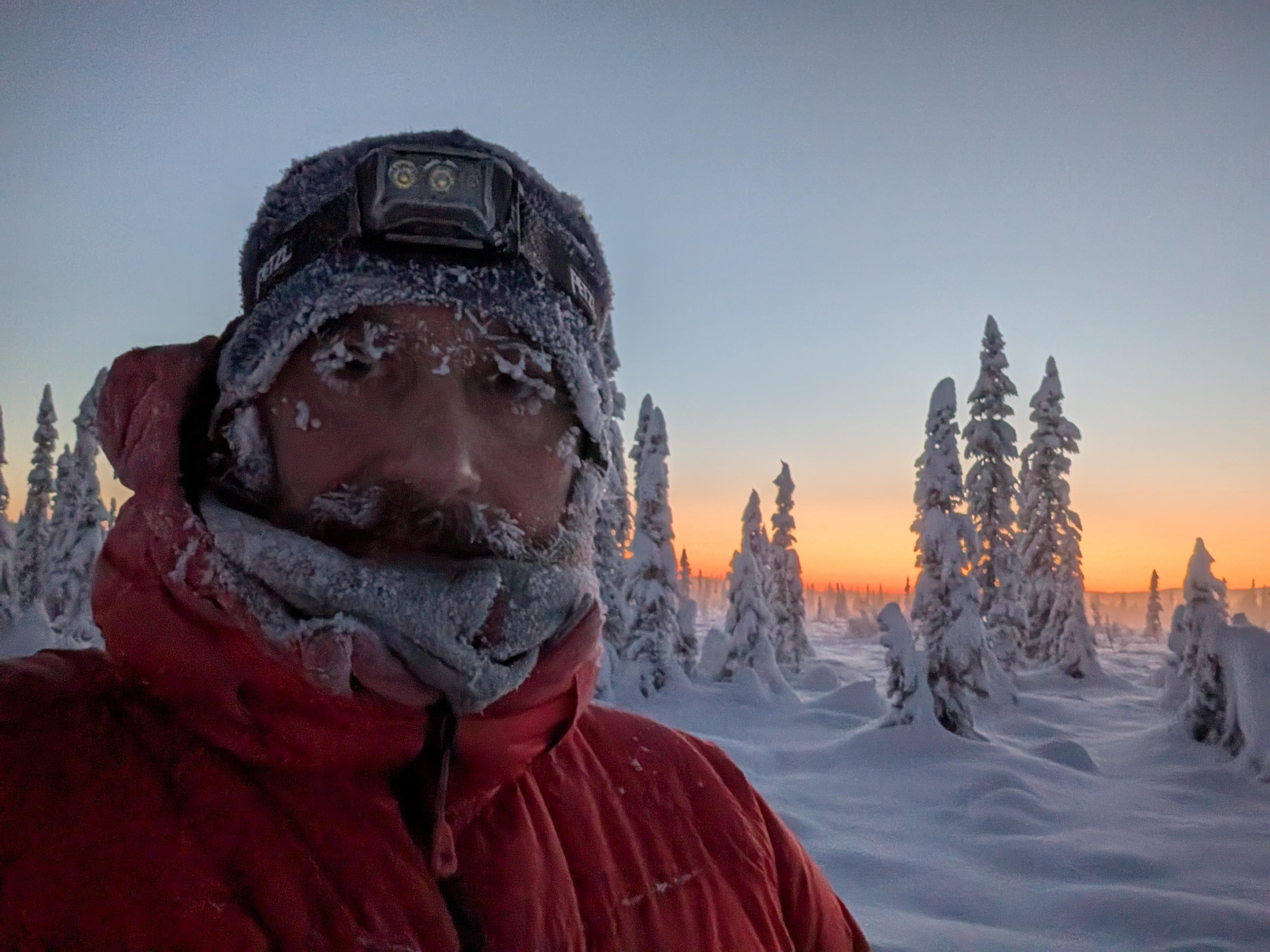 A close-up selfie of a person outdoors in extreme cold. The person is wearing a red insulated jacket, a winter hat, a scarf covering the lower face, and a headlamp on their forehead. Frost and ice cling to their eyebrows, eyelashes, and facial hair. Behind them, snow-covered spruce trees and a warm orange sunset glow are visible.