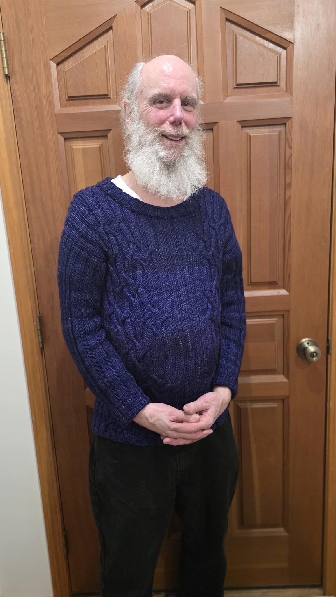 A smiling older man with a bushy white beard stands in front of a brown door. He's wearing a knit, cabled sweater in dark purple. It has a wide neck and long sleeves.