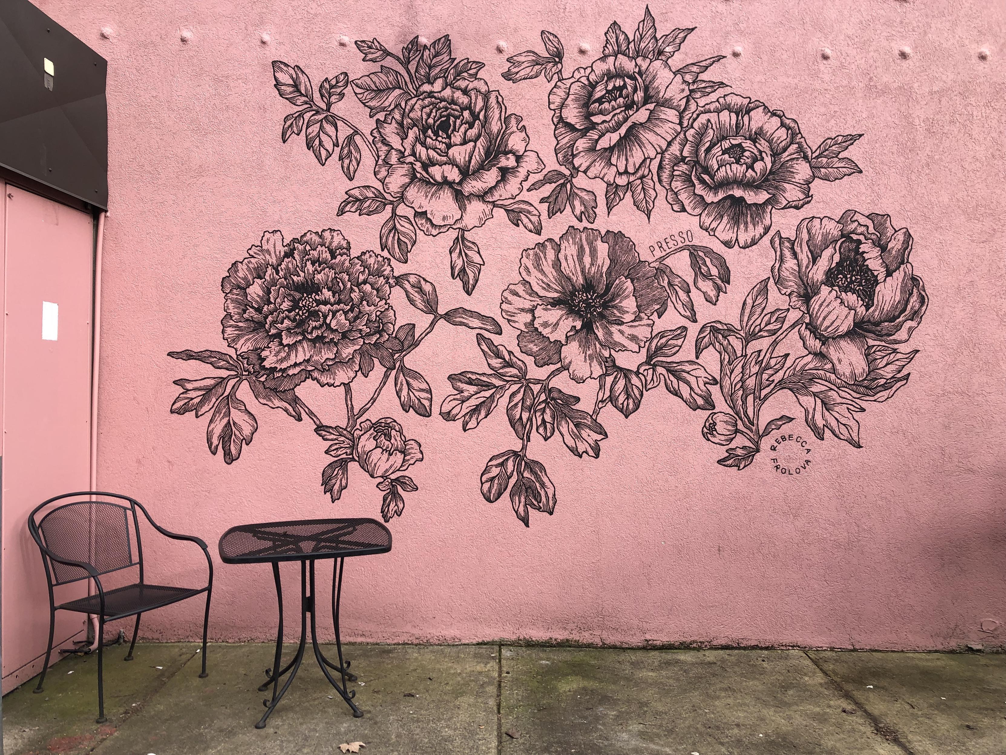 A line drawing mural of flowers on a pink wall with a little table on the sidewalk in front