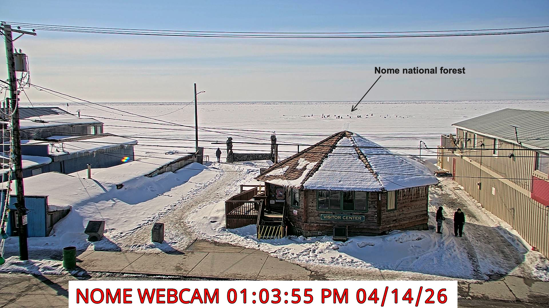 Webcam view looking south across Front Street in downtown Nome to Norton Sound beyond. The "Nome National Forecast" is visible on the sea ice a couple hundred yards offshore and blue skies with only a few clouds above.