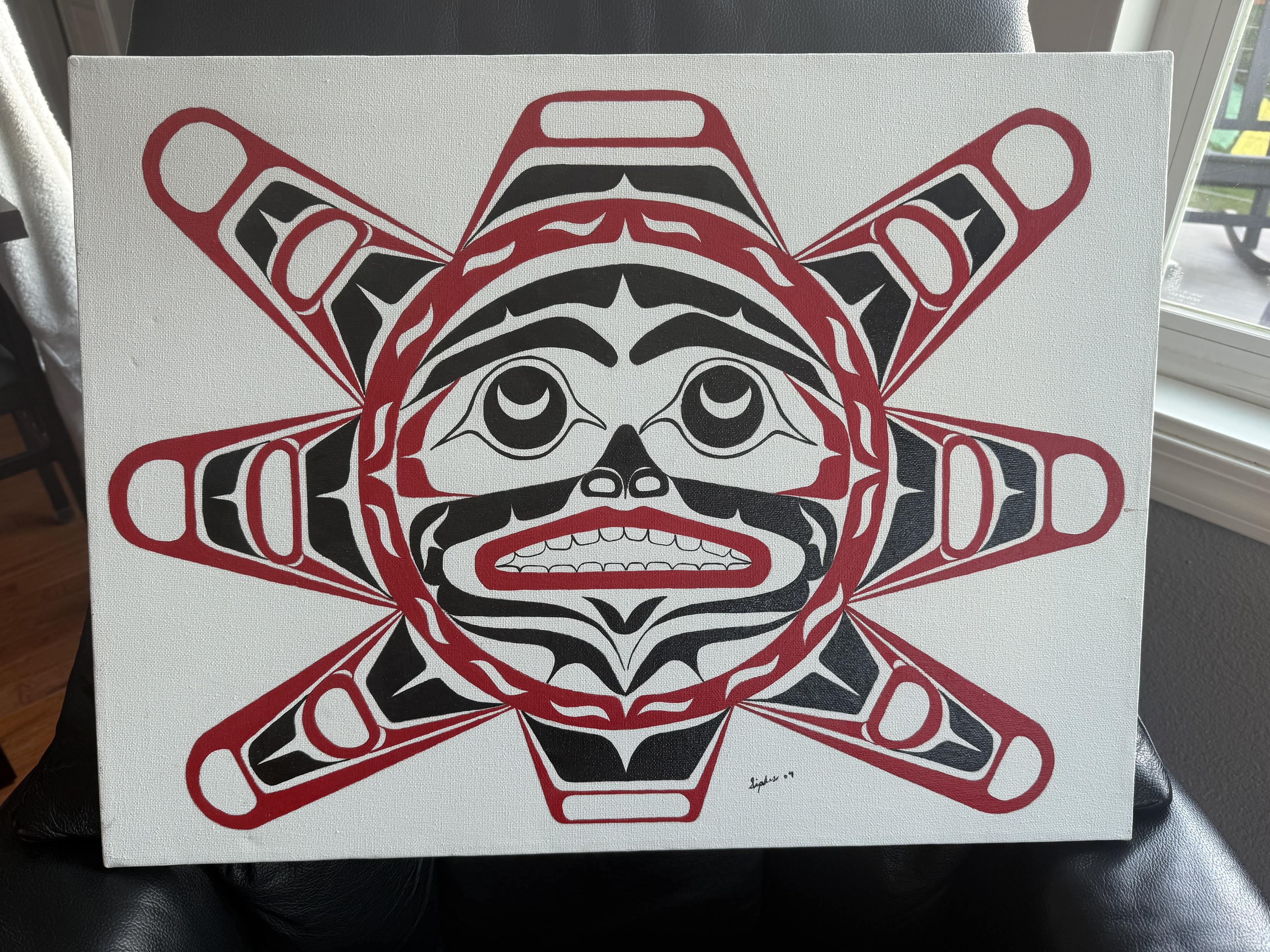 a Pacific Northwest Coast Indigenous formline sun by Alaska artist Paul Allen Siples (20th/21st Century). Using traditional colors of red and black, the central circle holds a face with wide, upturned eyes and an open mouth showing teeth, while eight symmetrical sun rays burst from all of its sides.