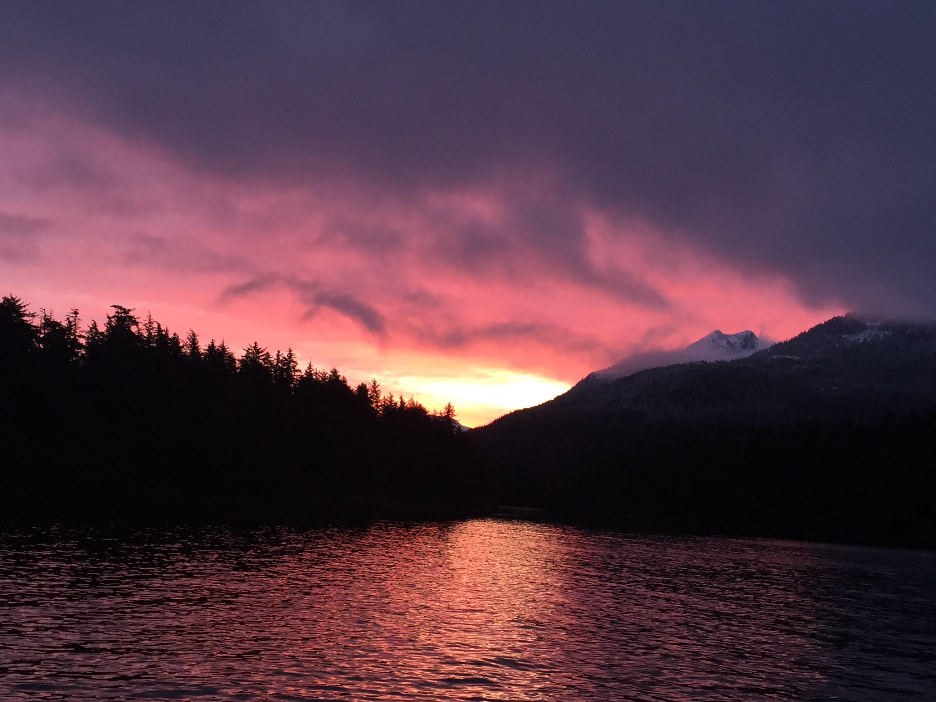 Deep purple and rust/red sunset between two islands that come together in a V. The bright yellow of the sinking sun is just visible below the red and dark purple clouds. The bottom 3rd is ocean, the subtly glows with orange/red.